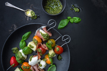 Apéro-Spiesse mit Tomaten, Käse, Schinken, Mozzarella und Pesto auf einem Teller auf einem alten dunklen Untergrund