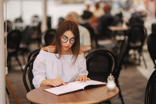 Stylish lady sits in cafe in centre of city and look at the menu. Sunny weather and codl drink. Girl in sunglasses chooses what to order