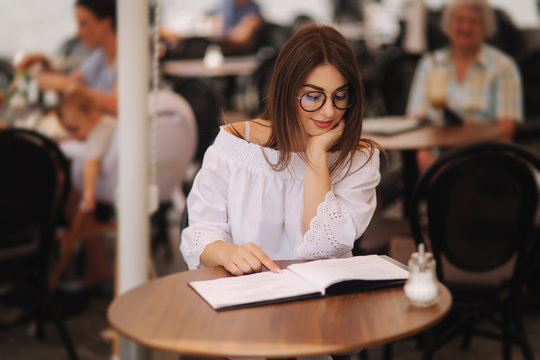 Stylish lady sits in cafe in centre of city and look at the menu. Sunny weather and codl drink. Girl in sunglasses chooses what to order
