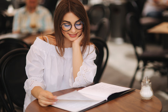 Stylish lady sits in cafe in centre of city and look at the menu. Sunny weather and codl drink. Girl in sunglasses chooses what to order