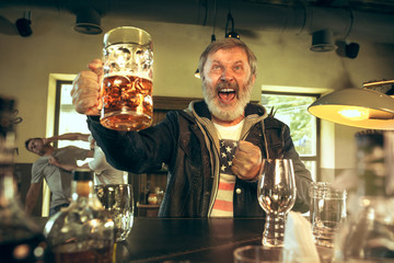 Senior bearded man drinking alcohol in pub and watching a sport program on TV. Enjoying beer. Man with mug of beer sitting at table. Football or sport fan. fight of fans in the background