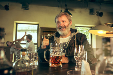 Senior bearded man drinking alcohol in pub and watching a sport program on TV. Enjoying beer. Man with mug of beer sitting at table. Football or sport fan. fight of fans in the background