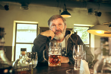 Sad senior bearded man drinking alcohol in pub and watching a sport program on TV. Enjoying my favorite teem and beer. Man with mug of beer sitting at table. Football or sport fan. Human emotions