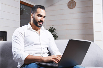 Handsome young enterpreneur, bearded man wears white shirt sitting on the sofa and working with...