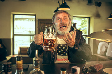 Sad senior bearded man drinking alcohol in pub and watching a sport program on TV. Enjoying my favorite teem and beer. Man with mug of beer sitting at table. Football or sport fan. Human emotions