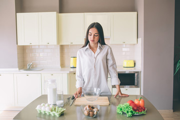 Serious and concentrated girl stands at table and look at products she has there. There are wood board and glass bowl. She is going to cook.