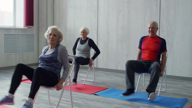 PAN of female aerobics instructor and cheerful senior people with grey hair doing chair fitness workout