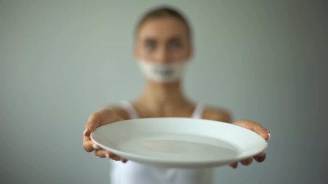 Skinny girl with taped mouth showing empty plate, concept of hunger and anorexia