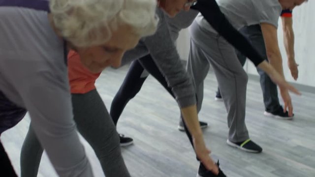 PAN with tilt up of elderly people in sportswear standing in line and trying to reach their toes while doing side bend exercise in yoga class