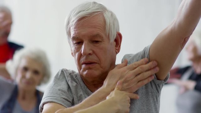 Medium shot of female fitness instructor positioning arms of senior man and explaining him how to do exercise during warm-up in group class; elderly people stretching in background