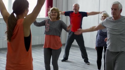 Tilt up of female fitness instructor and cheerful senior people in sportswear walking in place and doing dynamic stretching exercises during group class