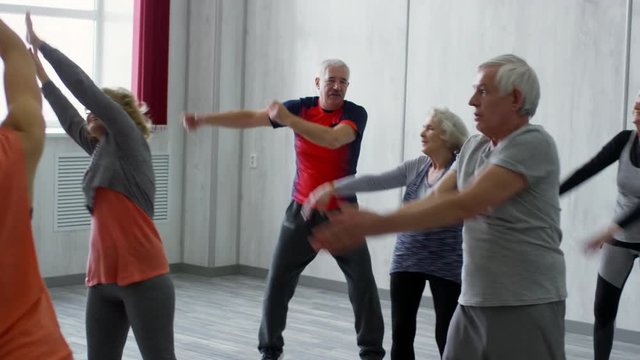 PAN of female fitness trainer and happy senior people in sportswear doing dynamic stretching exercises for arms during group class: they are swinging raised arms from side to side and smiling