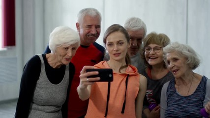 PAN of cheerful female fitness instructor and happy senior people with dumbbells laughing and taking selfie after class