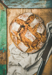 Flat-lay of freshly baked sourdough wheat bread loaf over linen napkin and rustic wooden table background, top view, vertical composition
