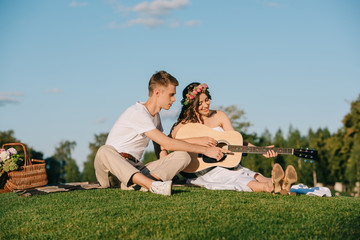 groom teaching bride to play on acoustic guitar during picnic with wicker basket