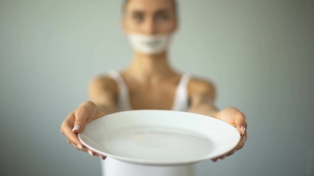 Slim girl with taped mouth showing empty plate, severe diet and self-destruction