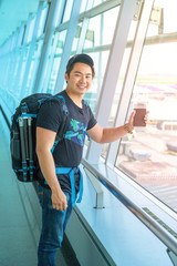 male tourist is standing in airport and looking at aircraft flight through window. He is holding tickets and backpack