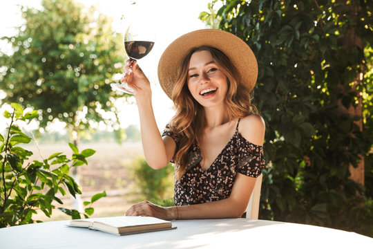 Photo of happy young woman wearing straw hat drinking wine, while sitting at table and reading book outdoor in cafe