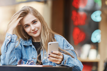 Beautiful dreamy european woman with fair hair in denim jacket, leaning on hand and touching strands, smiling with cute expression at camera, thinking how answer on message via smartphone