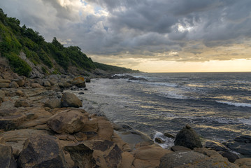A high rocky seashore at sunrise. Plants on the rocks. Waves and glare on the water.