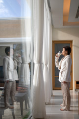 Asian woman drinking a glass of milk in living room and looking outside window with relaxed gesture