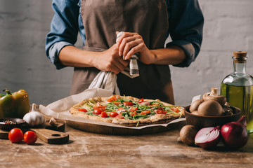 partial view of woman in apron standing at surface with cooked pizza and ingredients