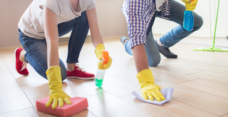 Girl and guy stand the floor with one hand. They wear yellow gloves. Girl holds sponge and spray...