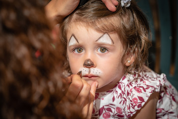 A cute 2 years old girl is being face-painted like a cat at a party