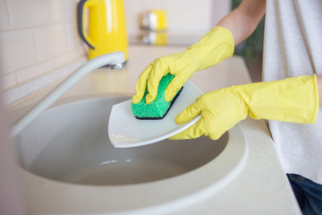 A picture of human's hands washing the dishes. People uses green sponge and wears yellow gloves.