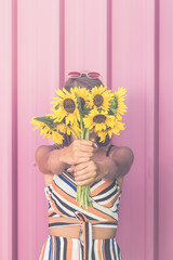 Hipster girl holding bouquet of sunflowers over her face on pastel pink background.