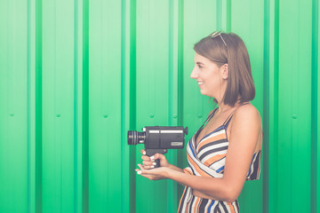 Young fashionable girl with vintage camcorder against green wall outdoors.