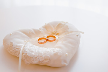 Two gold wide wedding rings on a white decorative pillow decorated with white lace and beads on a light background