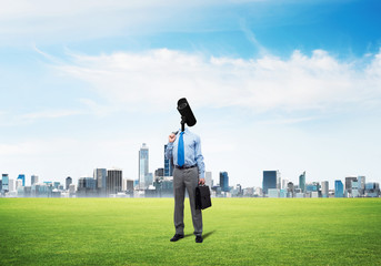 Camera headed man standing on green grass against modern cityscape