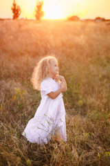 Fototapeta na wymiar Adorable happy little girl with a curly blonde hair, wearing a white dress , standing in the sunny sunset field among wild grass and flowers, smiling 