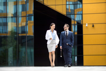 Young successful laughing male and female in business dress leaving office block, having dialogue