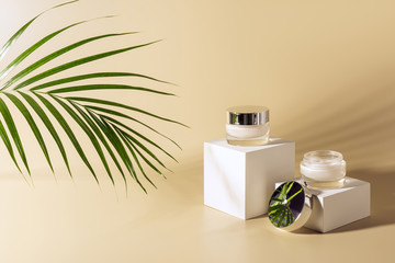 close up view of green palm leaf, facial and body creams in glass jars on white cubes on beige...