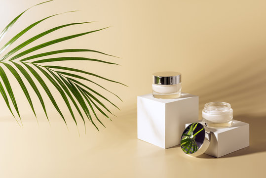 close up view of green palm leaf, facial and body creams in glass jars on white cubes on beige background