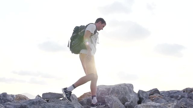 A tourist with a backpack behind him goes in sneakers on large stones high in the mountains, in the sunshine against the sky. slow motion. 1920x1080. full hd