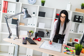 A young girl stands near a computer desk and draws a marker on a magnetic board.