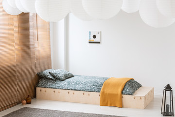 Orange blanket on wooden bed in white bedroom interior with lantern, poster and blinds. Real photo
