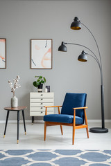 Flowers on wooden table next to blue armchair and lamp in grey living room interior. Real photo