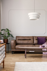 Real photo of modern lamp hanging above leather couch with purple pillow in bright living room...