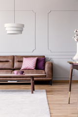 Real photo of pink and purple pillows placed on leather couch in bright living room interior with wainscoting on wall, coffee table with two cups and modern lamp