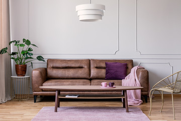 Real photo of brown leather sofa with violet cushion and pastel pink blanket standing in light grey...