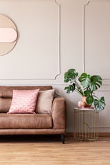 Real photo of end table with decor and fresh plant placed next to leather sofa with pink cushions in light grey living room interior with wainscoting on wall