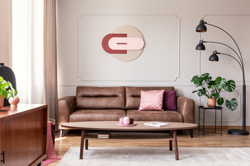 Real photo of wooden coffee table placed on carpet in bright sitting room interior with leather brown couch, modern clock on wall, metal lamp and window with drapes - obrazy, fototapety, plakaty