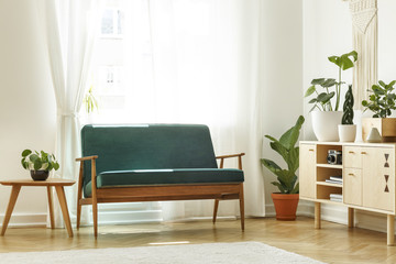 Real photo of a retro sofa next to a coffee table and cabinet with plants and window in the...