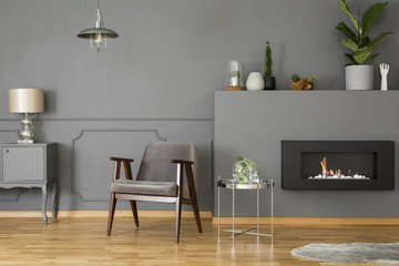 Grey armchair next to silver table in elegant living room interior with fireplace and lamp. Real...