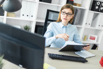 A young girl in glasses sits at a table in the office, holds a pencil in her hand and works with documents.