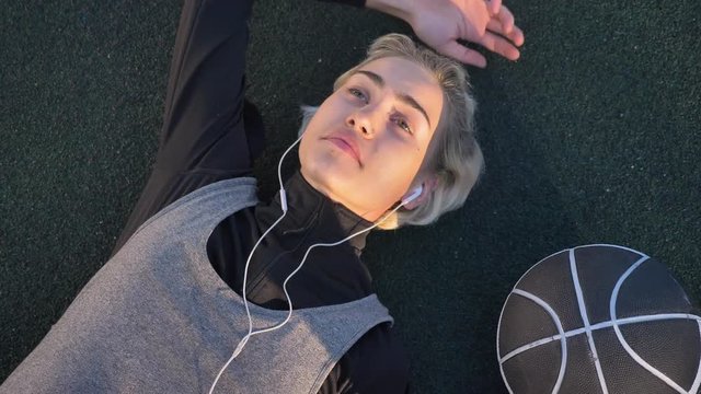 Dreamy young female basketball player lying on court and listening music through earphones, ball placing near her, peaceful and calm